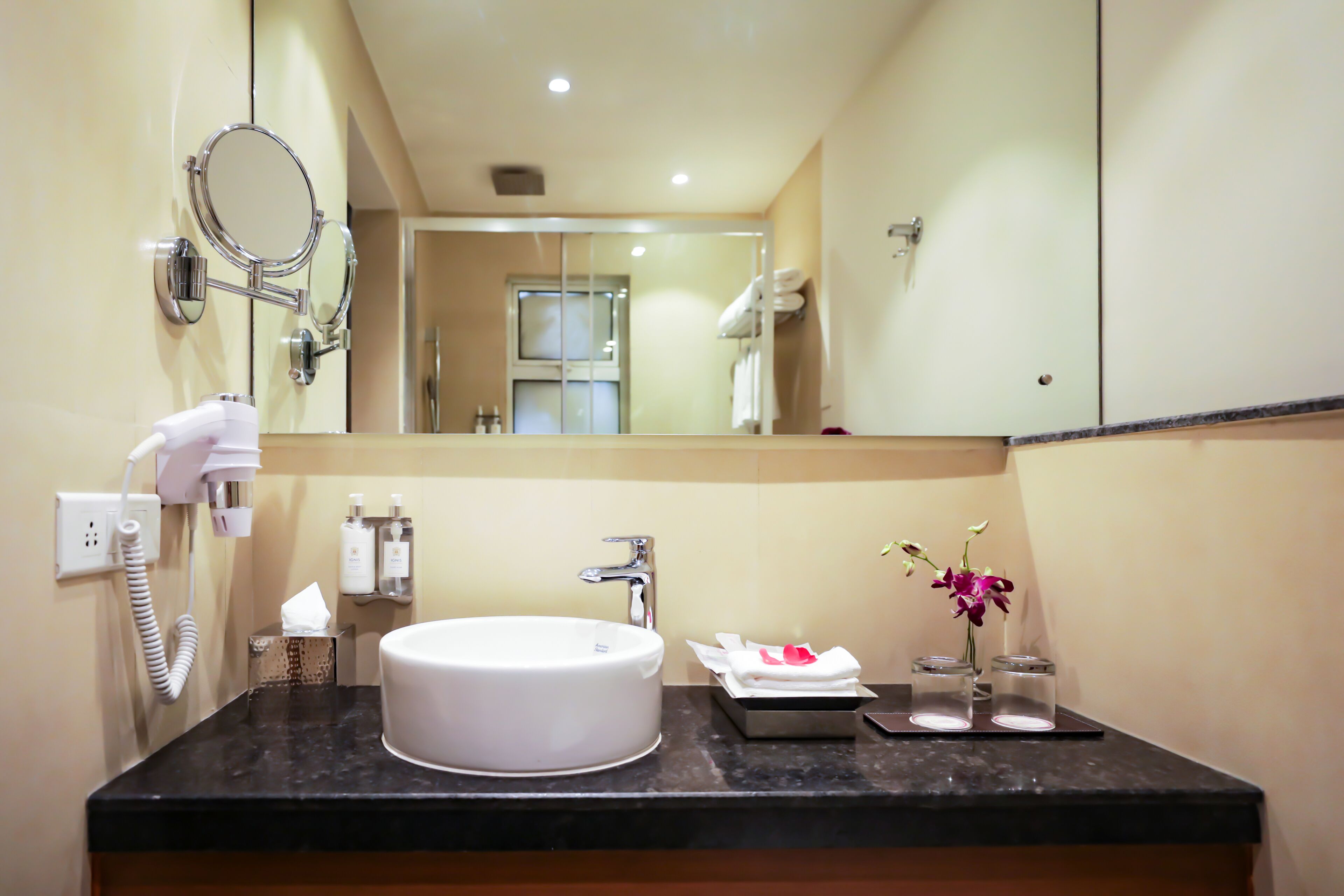 A bathroom with a round sink, a mirror, a hairdryer, and a vase with flowers.