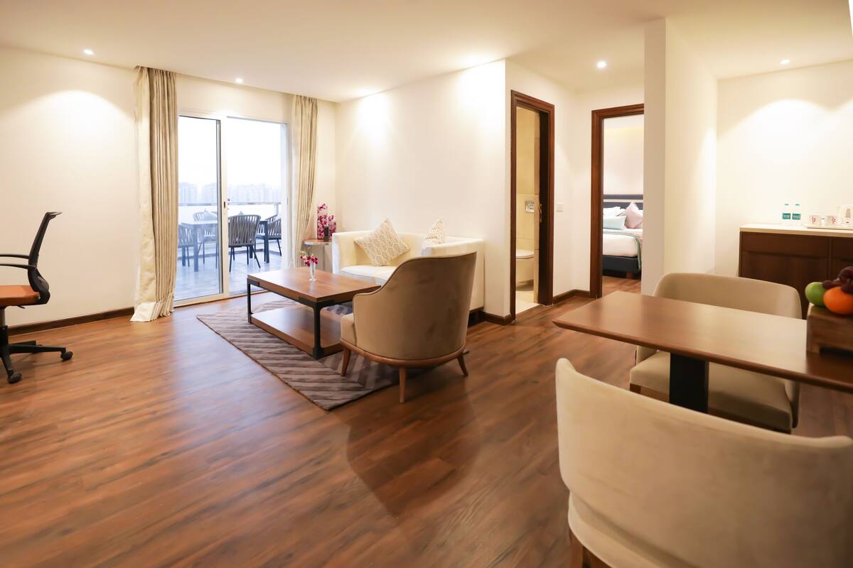 A modern hotel room with a wooden floor, a dining area with a table and chairs, a sofa, and a view through a large window.
