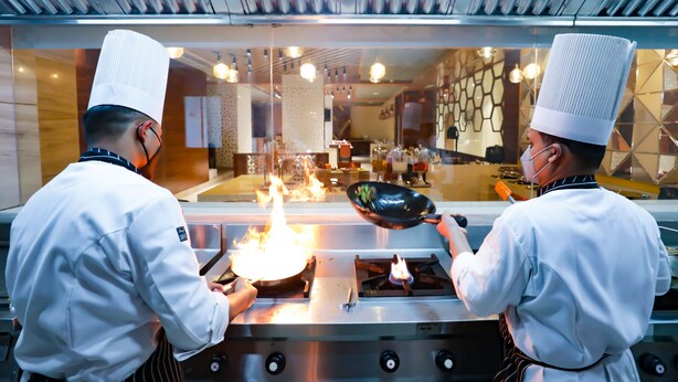 Dos cocineros con uniformes blancos y gorros altos cocinando en una estufa con llamas.