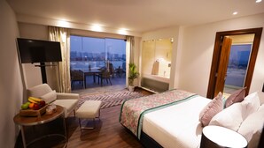 Habitación de hotel con una cama grande, televisión, zona de comedor con sillas y una mesa, y vistas a la ciudad.