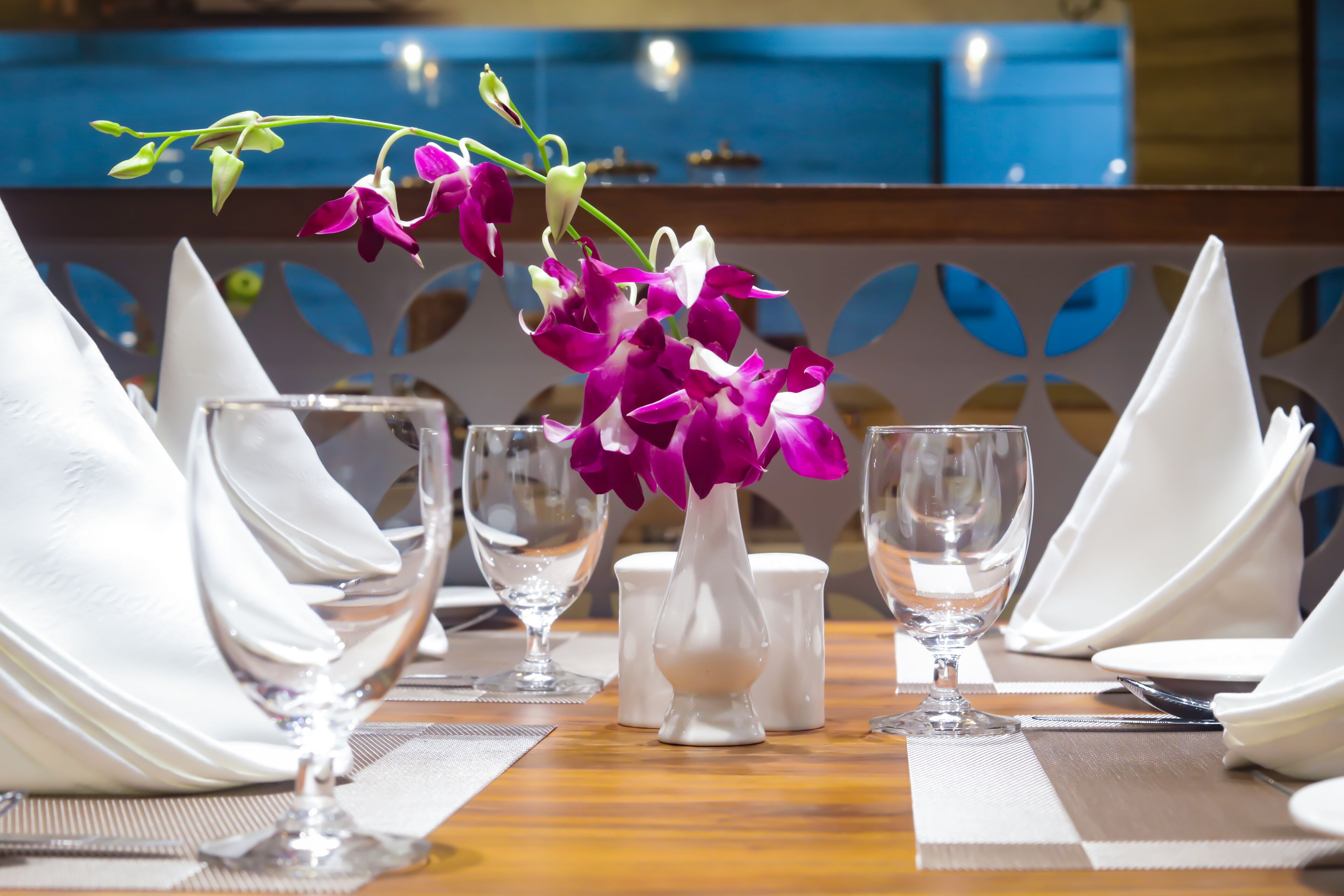 Una mesa dispuesta para comer, con servilletas blancas, vasos transparentes y un jarrón de orquídeas púrpuras.
