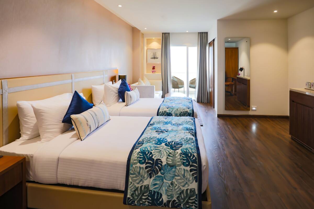 A hotel room with two beds, a wooden floor, a balcony with a chair, and a desk with a chair.