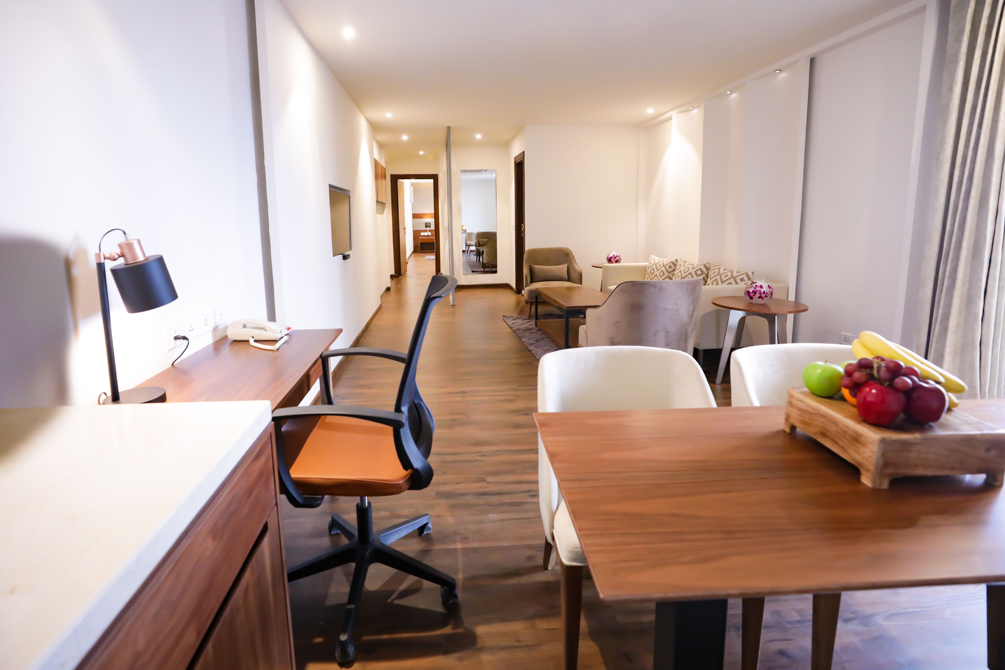 A hotel room with a wooden desk, office chair, and a small table with fruit. There is a sofa and a chair in the living area.