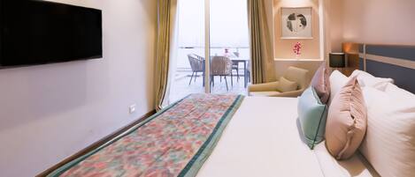 A hotel room with a bed, a TV mounted on the wall, a balcony with a table and chairs, and a framed picture on the wall.