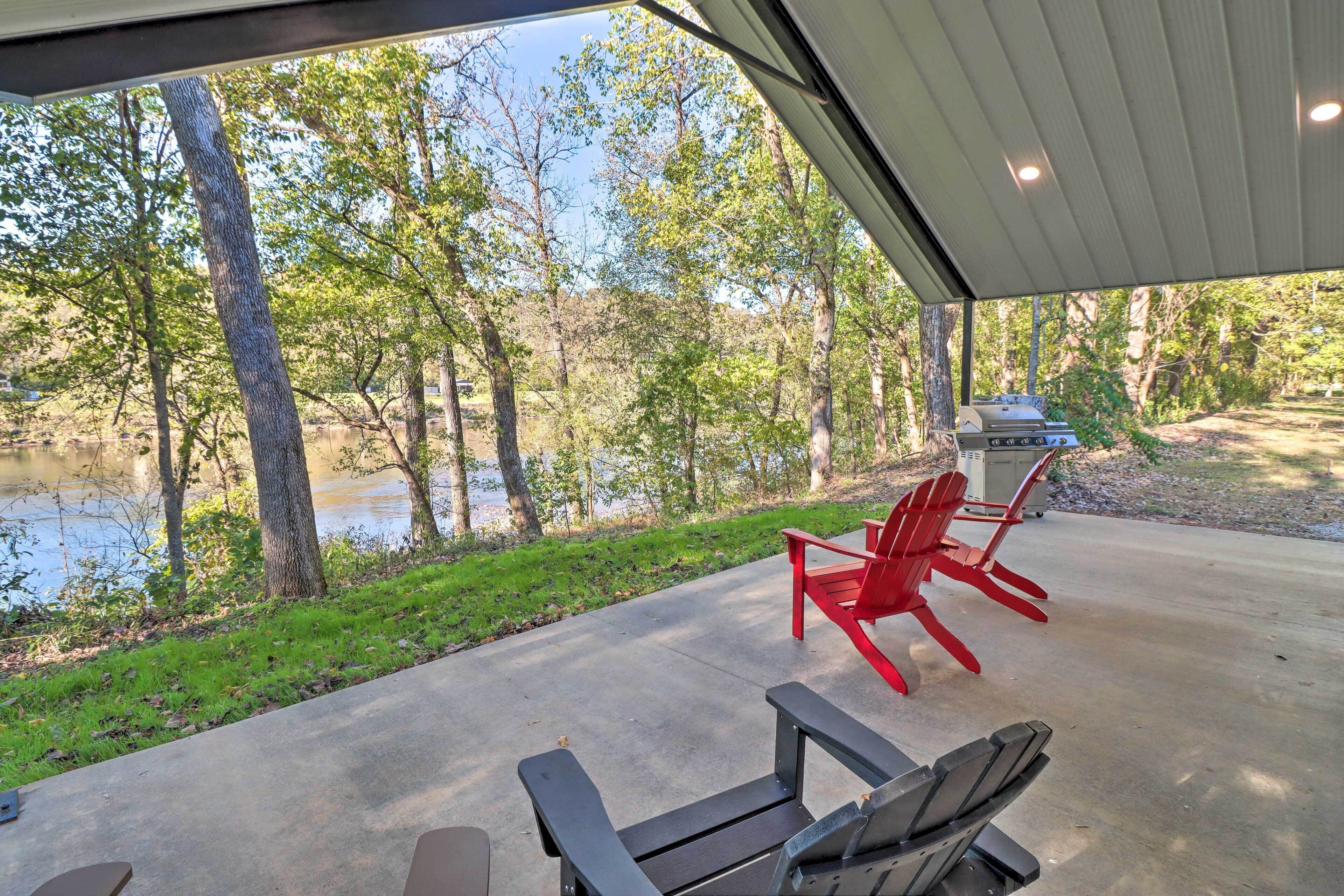 A covered patio with a view of a lake and trees, featuring two Adirondack chairs and a grill.