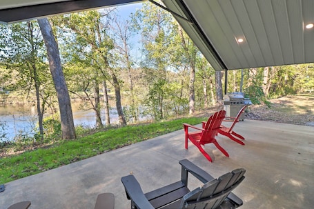 A covered patio with a view of a lake and trees, featuring two Adirondack chairs and a grill.