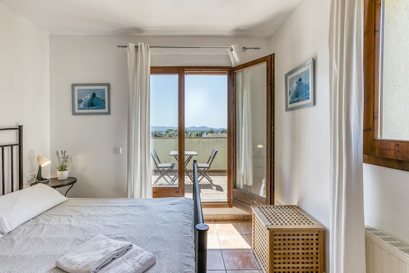 A bedroom with a bed, a nightstand, a potted plant, a window with curtains, and a balcony with a table and chairs.