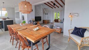 Un grand espace à manger avec une table et des chaises en bois, une cuisine en arrière-plan et un canapé en osier agrémenté d’un coussin.