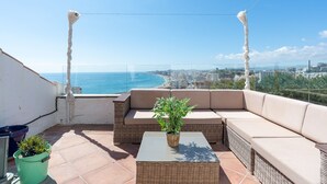 A balcony with a wicker sofa, a small table with a plant, and a view of the sea and cityscape.