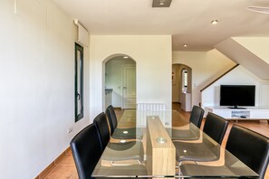 Uma área de jantar moderna com mesa de vidro, cadeiras pretas e uma TV ao fundo.
