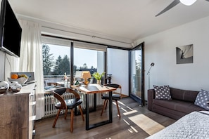 A modern living room with a sofa, a dining table with chairs, a TV, and a view through large glass doors.
