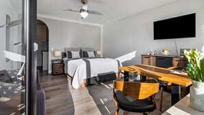 A modern hotel room with a bed, desk, chairs, a television, and a ceiling fan.