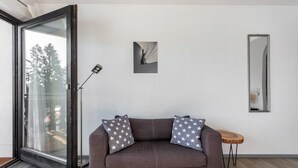 A modern living room with a brown sofa, two pillows with star patterns, a small round wooden side table, a floor lamp, a framed picture on the wall, and a large mirror.