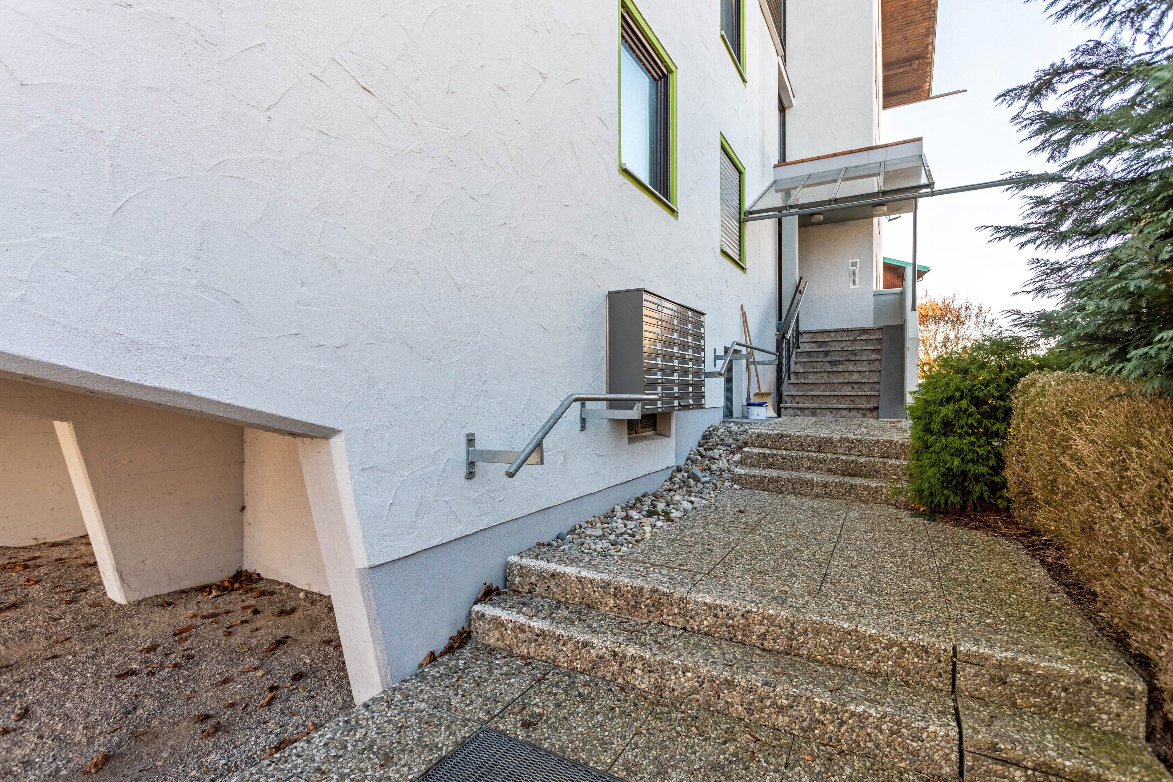 Ein Gebäude mit heller Fassade, einer Treppe aus Stein zum Eingang und einem kleinen Baum zur Rechten.