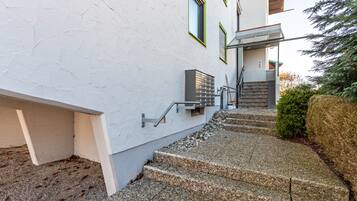 Ein Gebäude mit heller Fassade, einer Treppe aus Stein zum Eingang und einem kleinen Baum zur Rechten.