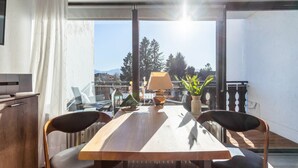 A dining area with a table, chairs, a lamp, and a view of trees and mountains through large windows.