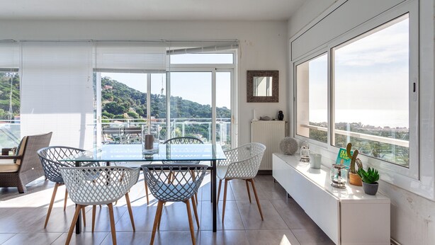 Un comedor moderno con una mesa de vidrio y sillas de metal, una consola blanca con objetos decorativos y ventanas grandes que ofrecen vistas a una ladera.