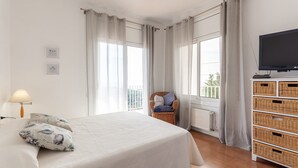 Un dormitorio con cama, televisión, un mueble de almacenamiento de mimbre y una ventana con cortinas.
