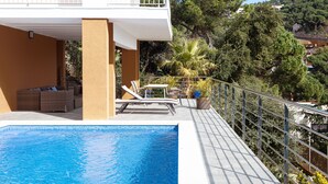 Zona de piscina con sillones de descanso, una mesita y vista a un cerro con árboles y edificios.
