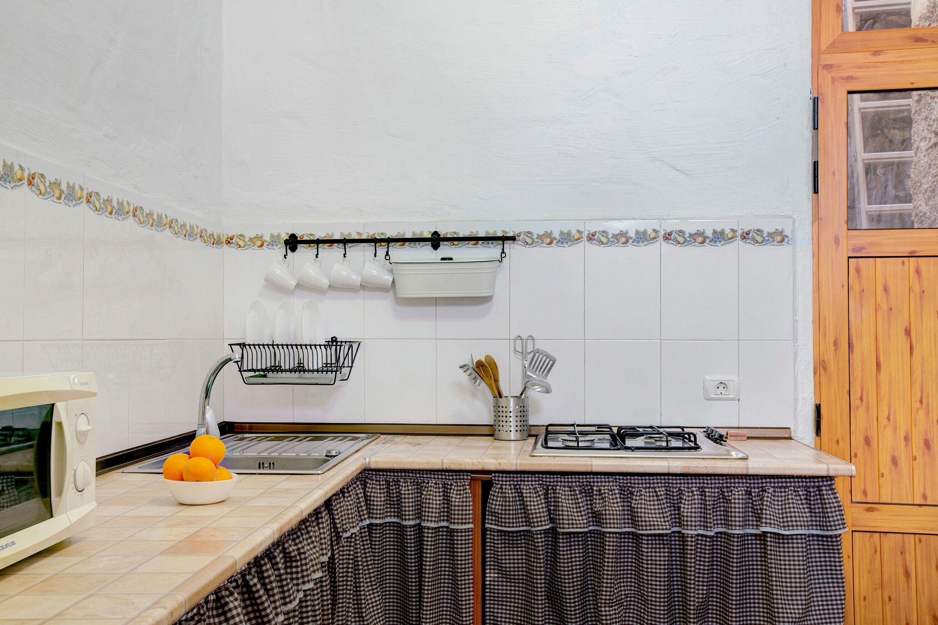 Una cocina con microondas, fregadero y estufa. Hay naranjas en la encimera, una cesta y un colgador con tazas.
