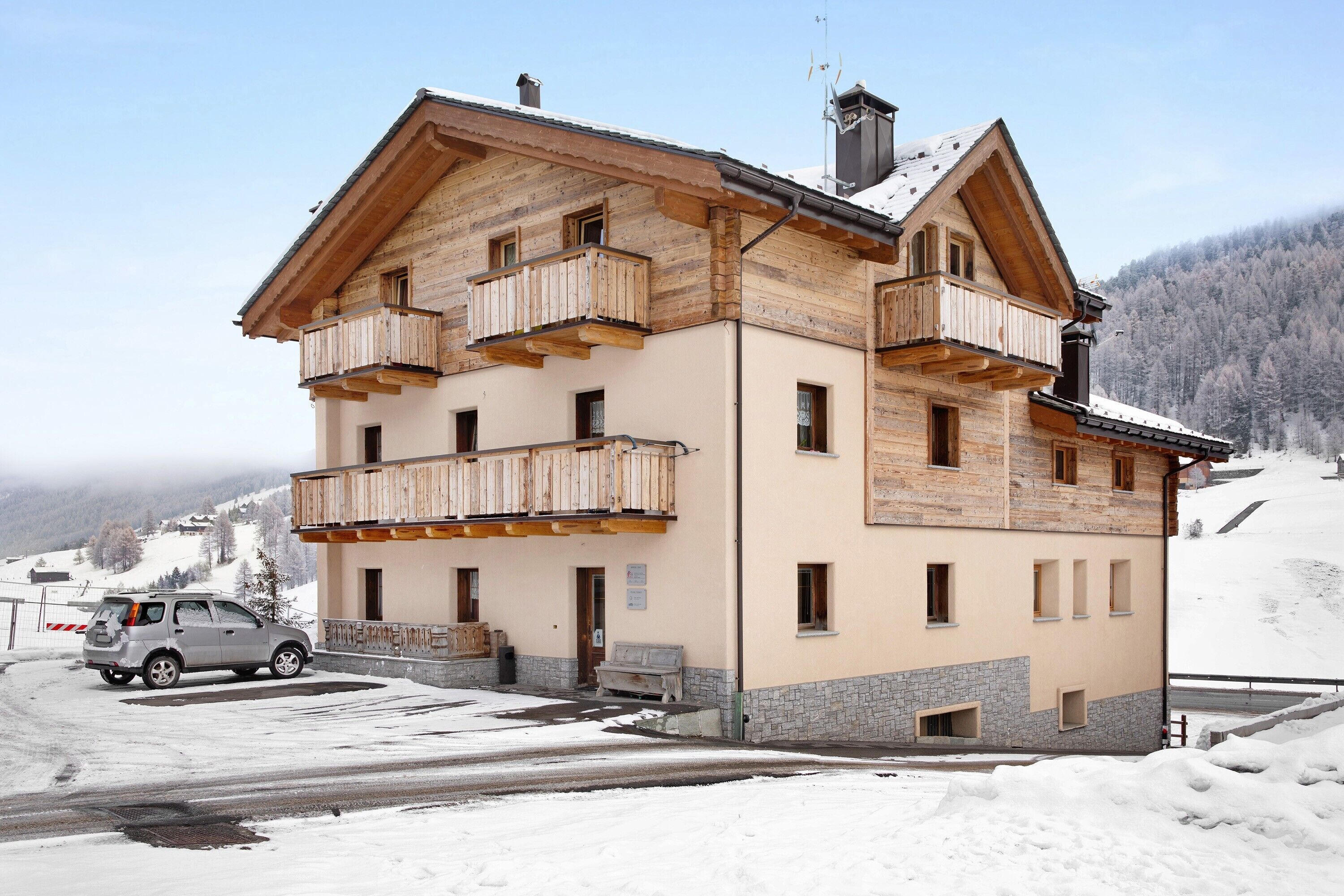 A multi-story building with balconies, a car parked in front, and a snowy landscape in the background.