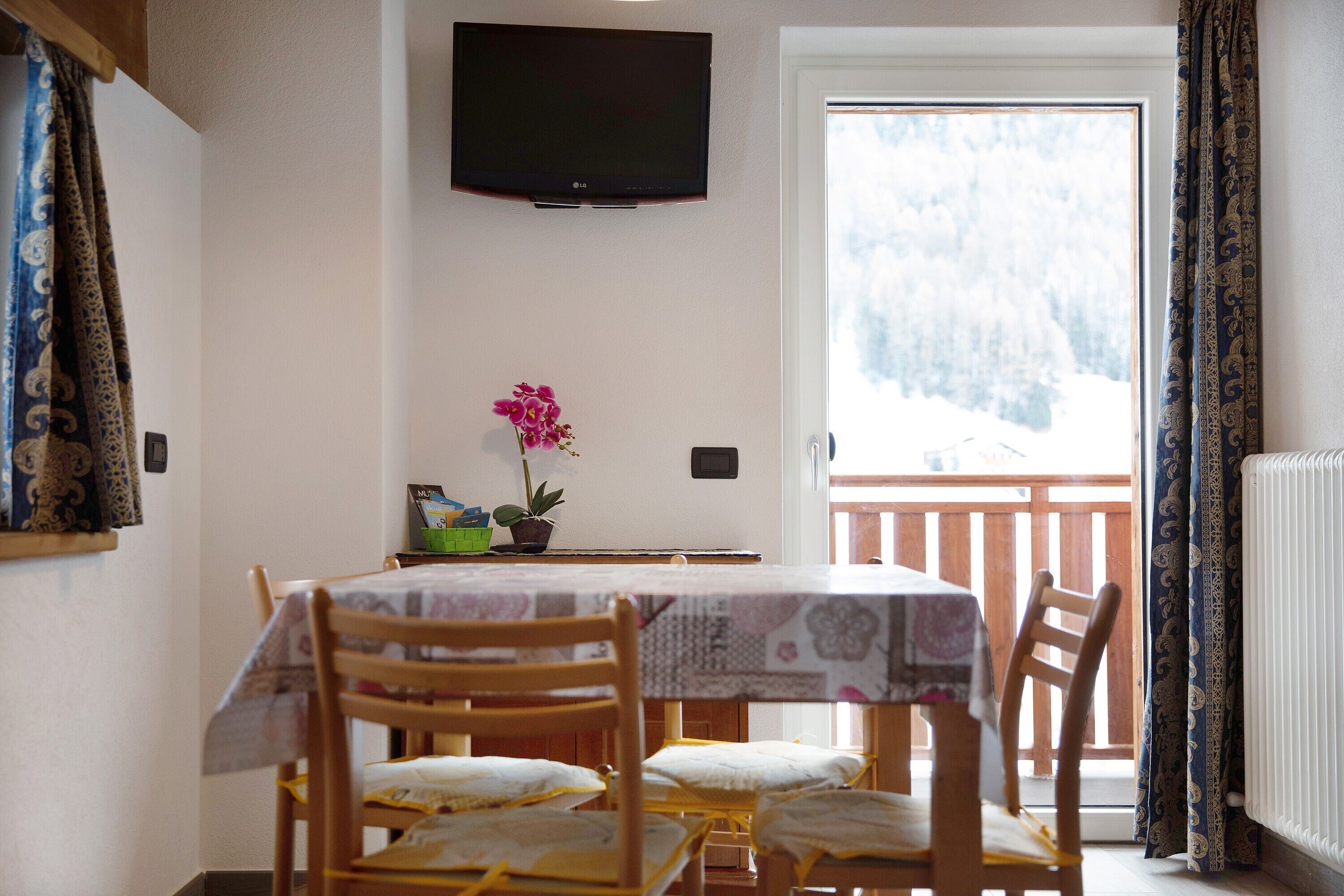 Un coin repas avec une table et des chaises, un vase de fleurs, un téléviseur fixé au mur et une vue sur un paysage enneigé par la porte.