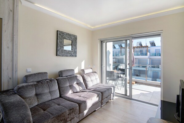A modern living room with a grey sofa, a framed artwork, and a sliding glass door leading to an outdoor balcony.