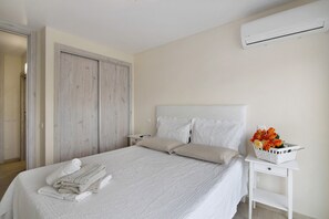 Un lit bien fait, avec du linge de lit blanc et des oreillers, une table de chevet sur laquelle se trouve un panier de fleurs, et un appareil de climatisation fixé au mur.