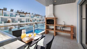 Eine Dachterrasse mit Glasbrüstung, einem Backstein-Grill, einem Esstisch mit Frühstücksgebäck und Blick auf einen Pool.
