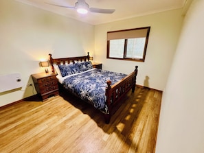 A bedroom with a wooden bed, a nightstand, a lamp, a ceiling fan, and a window with blinds. - Treetops Hideaway*Hikes*Close to Town*Nature (West Albury)