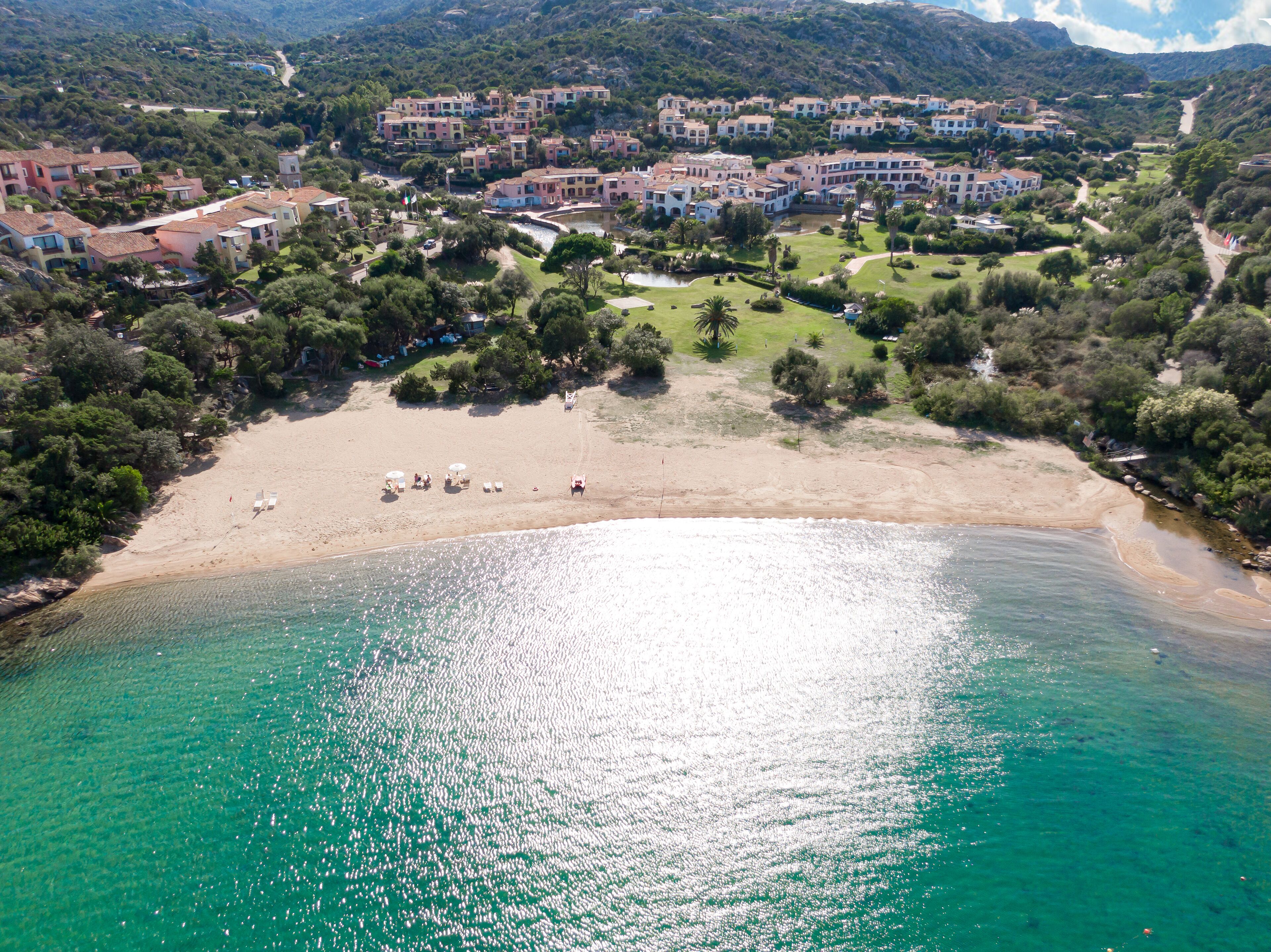 An aerial view of a coastal area with a sandy beach, clear turquoise waters, and a cluster of buildings with balconies overlooking the beach.