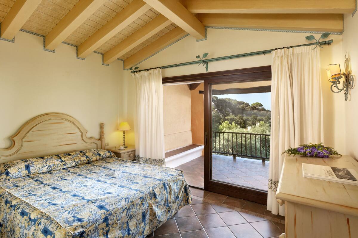 Un dormitorio con cama, mesita de noche, lámpara, balcón con vistas al exterior y ventana con cortinas.