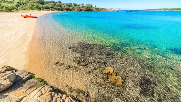 Una playa con agua turquesa cristalina, un bote en la arena y un litoral rocoso.
