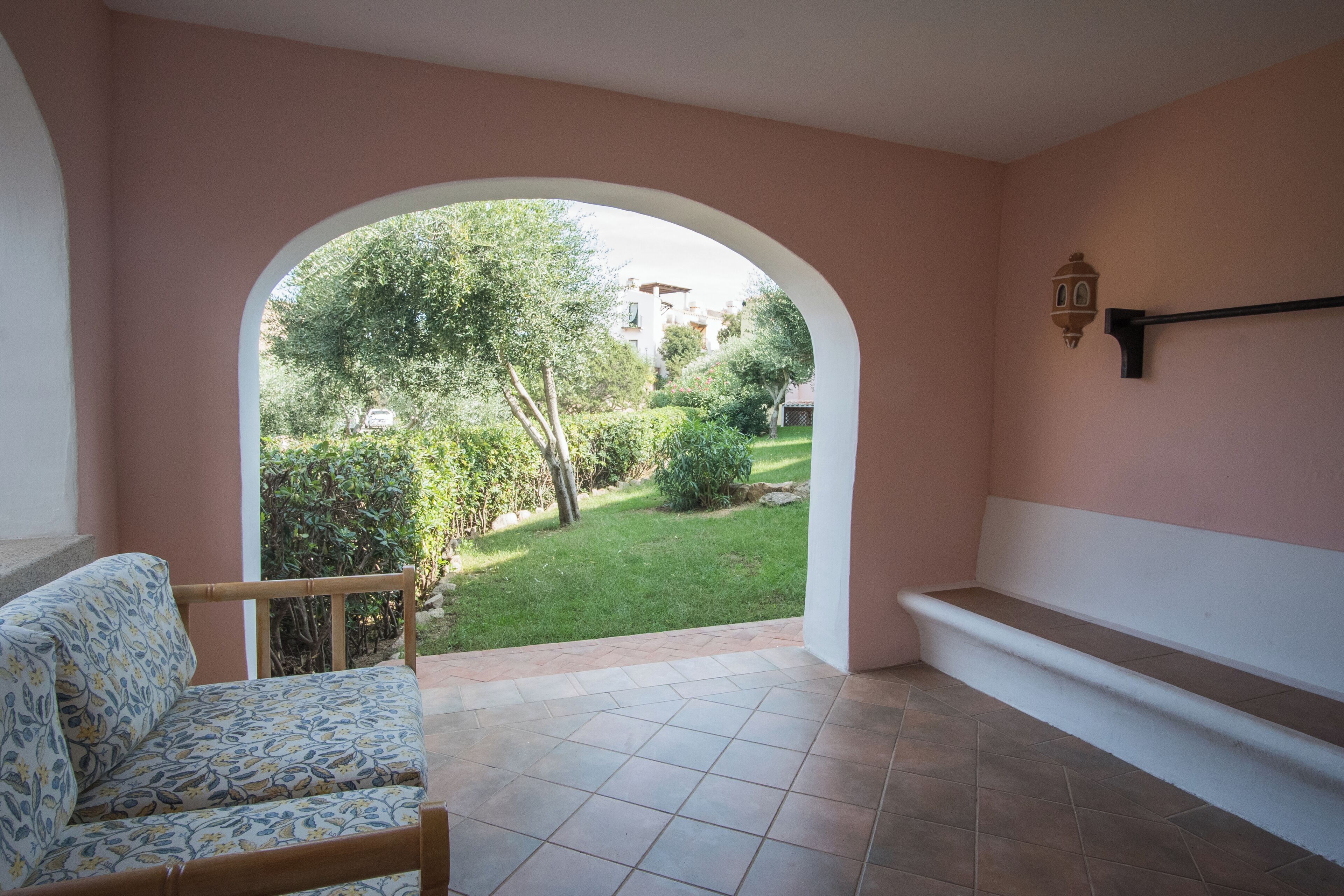 A room with a patterned sofa, a tiled floor, a bench, and an arched doorway leading to a garden with trees and a building in the background.