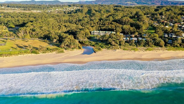 Una vista aerea di una zona costiera con spiaggia, un piccolo lago e un’area residenziale con case e vegetazione.