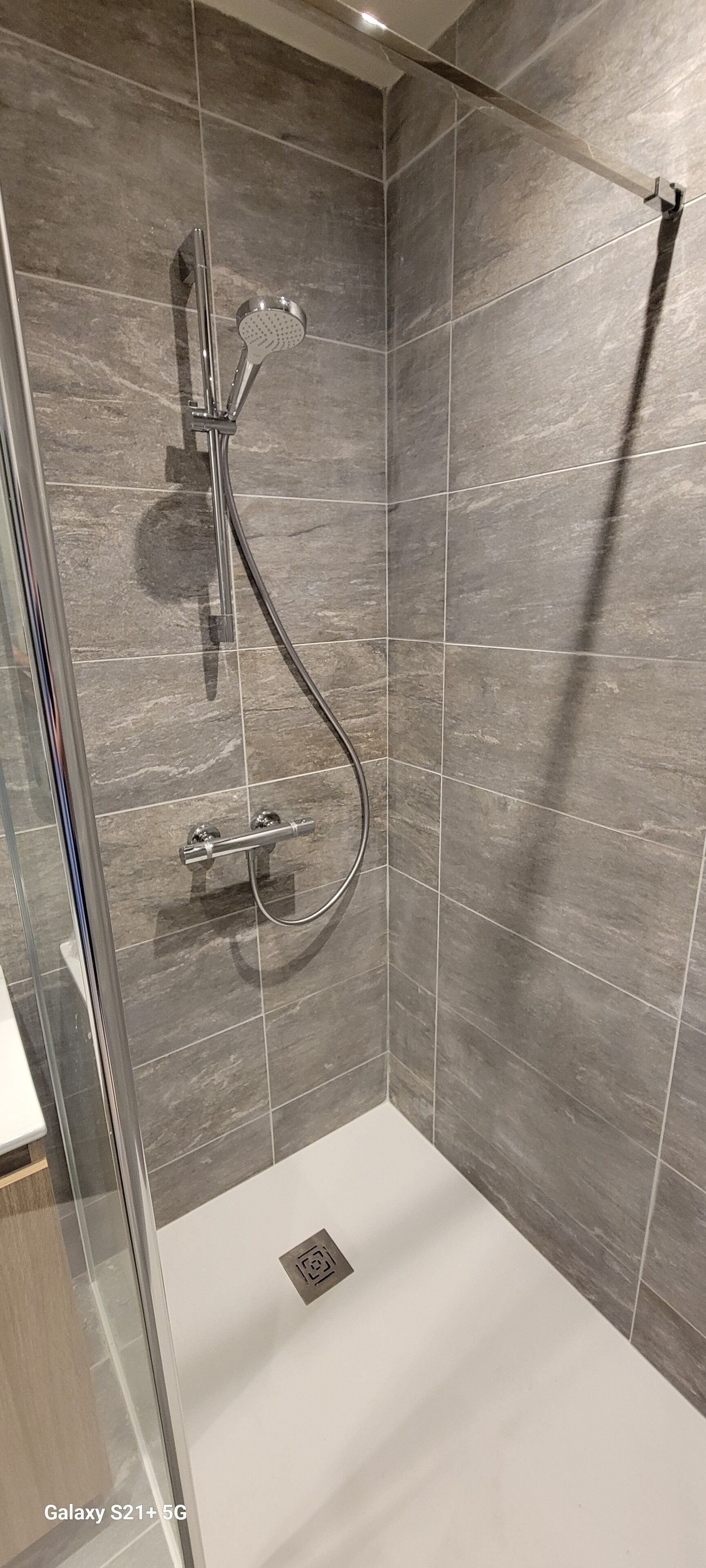 Een moderne douche met een glazen deur, grijze tegels en een handdouche.
