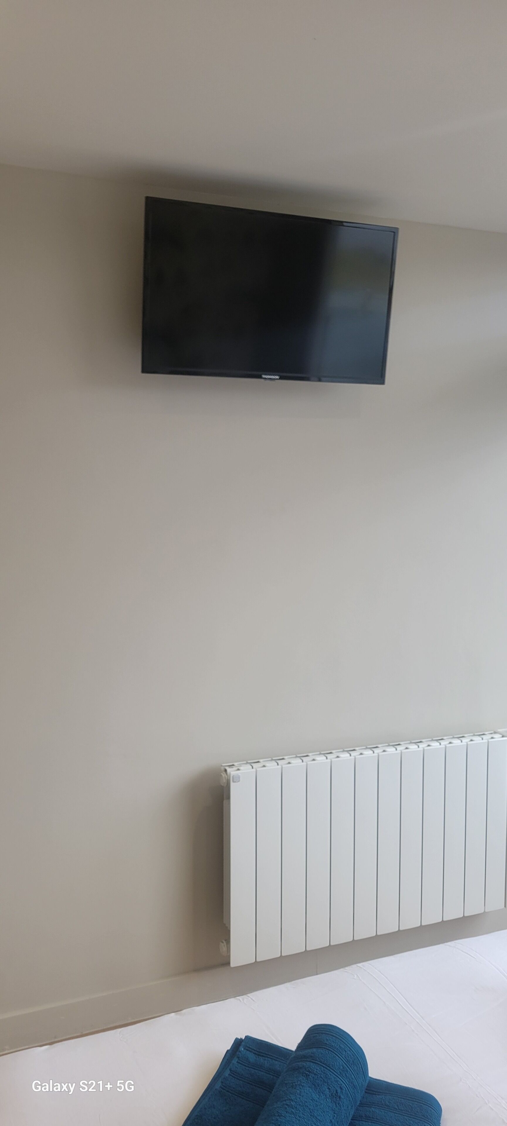 Ein Zimmer mit einem an der Wand befestigten Flachbildfernseher, einem weißen Heizkörper und einem Bett mit blauer Bettwäsche.