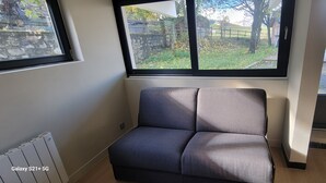 Ein Eckzimmer mit grauem Sofa, Heizkörper und Fenster mit Blick auf Garten und Gebäude.