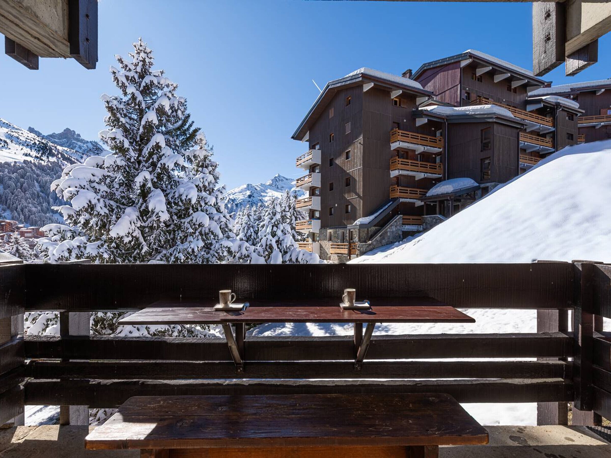 A wooden bench with a table, overlooking a snowy mountain landscape and a building with balconies.