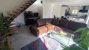 A modern living room with a brown leather sectional sofa, a glass coffee table, and a piano in the background. - Vacation home rental Gulf of Morbihan (Sarzeau)