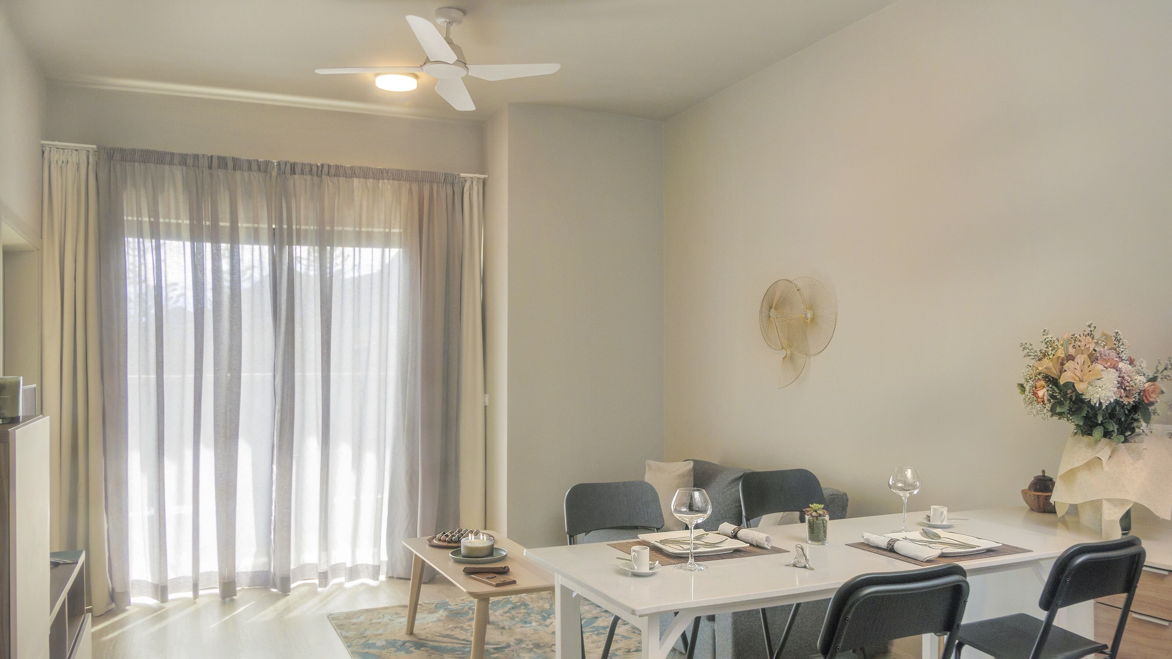 A dining area with a table set for four, a ceiling fan, and a large window with curtains.