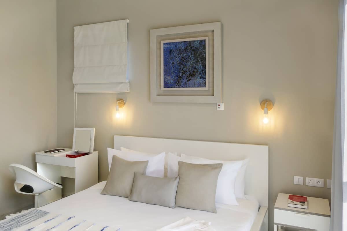 Una habitación de hotel con cama, mesita de noche, escritorio con silla, lámpara de pared y un cuadro enmarcado.