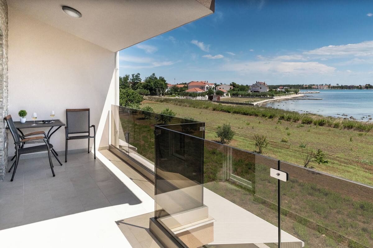 A balcony with a table and chairs, a glass railing, and a view of a coastal area with houses and greenery.