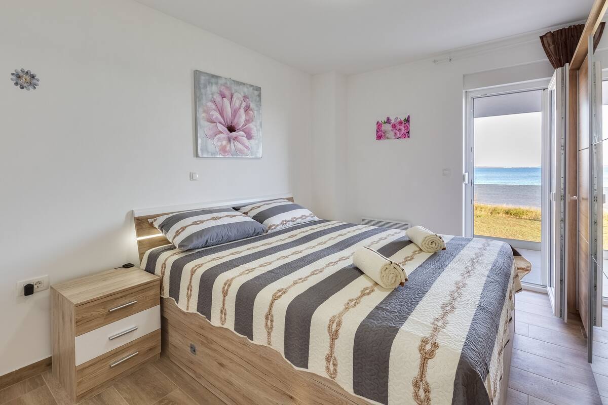 A bedroom with a bed, a nightstand, a large window with a beach view, and a wall with a flower painting.