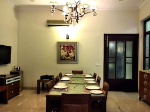 A dining room with a long table set for six, a chandelier, an air conditioner, a painting, and a television on the wall.
