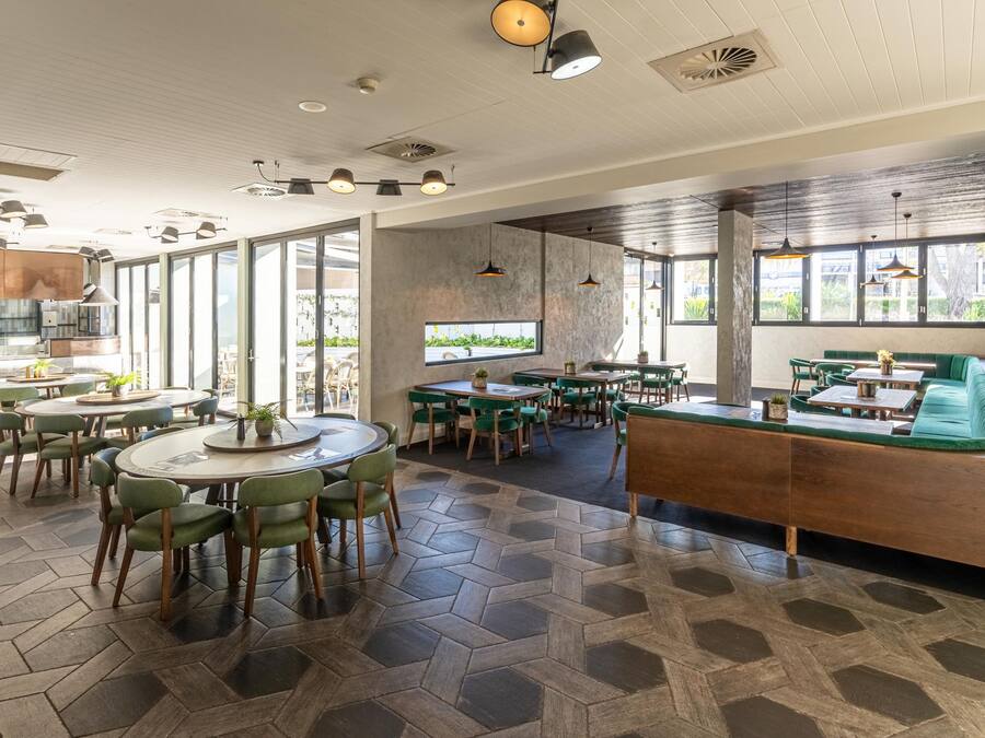 A modern restaurant with round tables, green chairs, and a central seating area with a green sofa and a wooden bench.