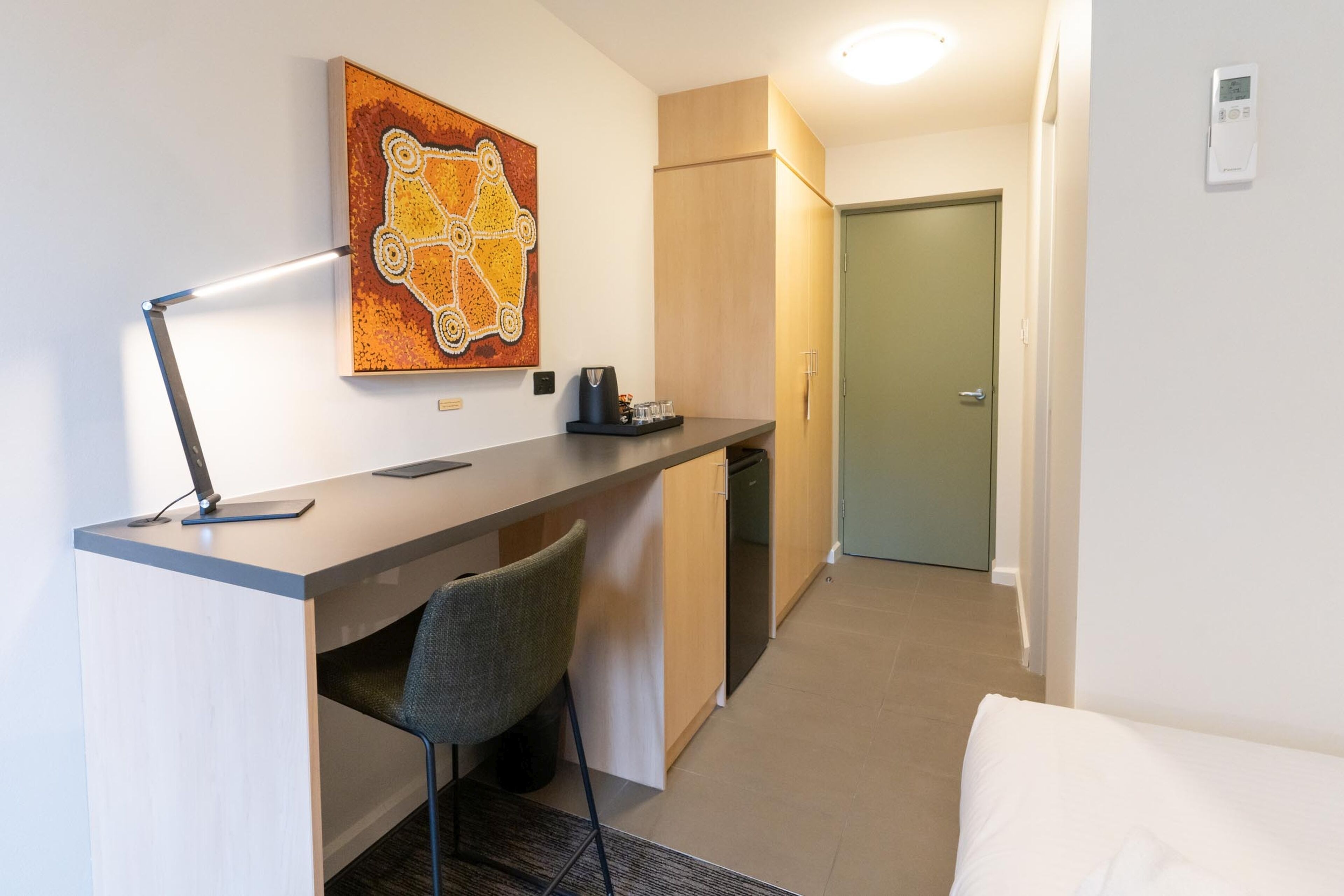 A compact hotel room with a kitchenette, a desk with a chair, a bed, and a painting on the wall.