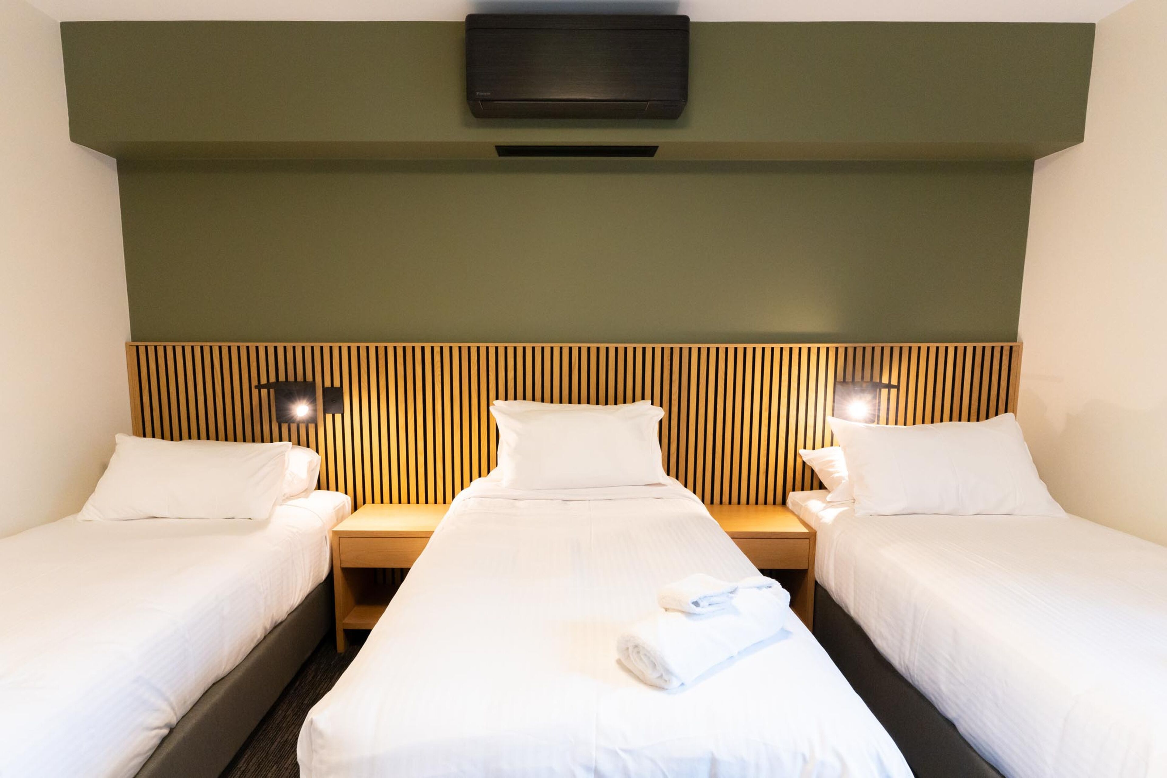Una habitación de hotel con dos camas individuales, cabecera de madera, mesitas de noche con lámparas y aire acondicionado instalado en la pared.