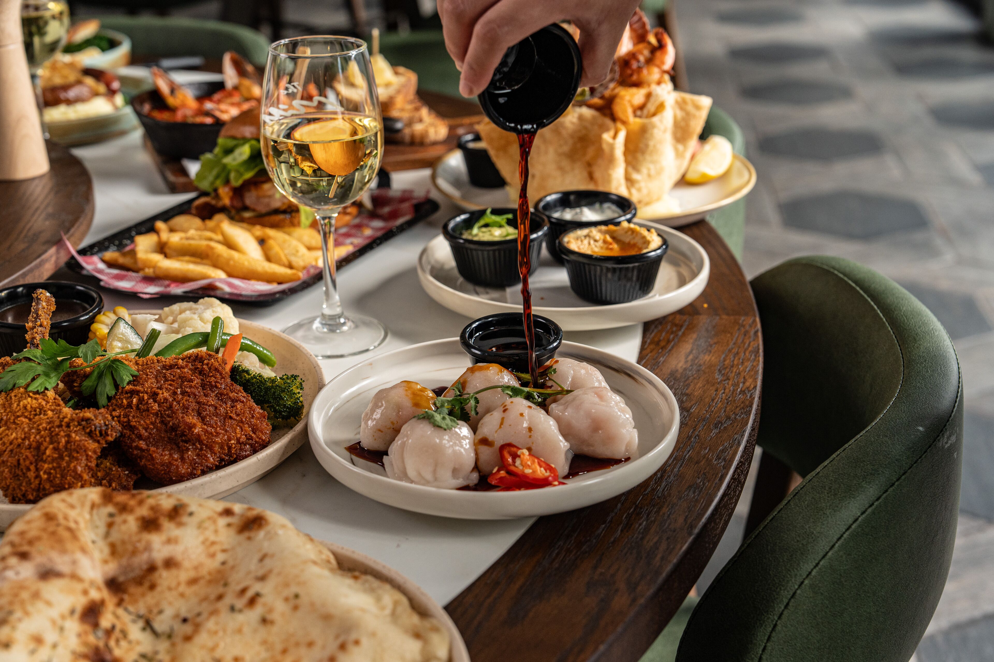 Una mesa con varios platos, incluyendo preparaciones fritas, una copa de vino blanco y una persona sirviendo salsa.