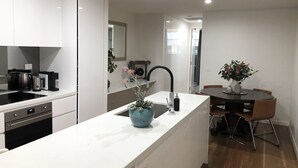 Eine moderne Küche mit weißen Schränken, einem eingebauten Backofen und einer Kaffeemaschine. Es gibt eine kleine Essecke mit Tisch und Stühlen, und eine Topfpflanze steht auf der Arbeitsfläche.